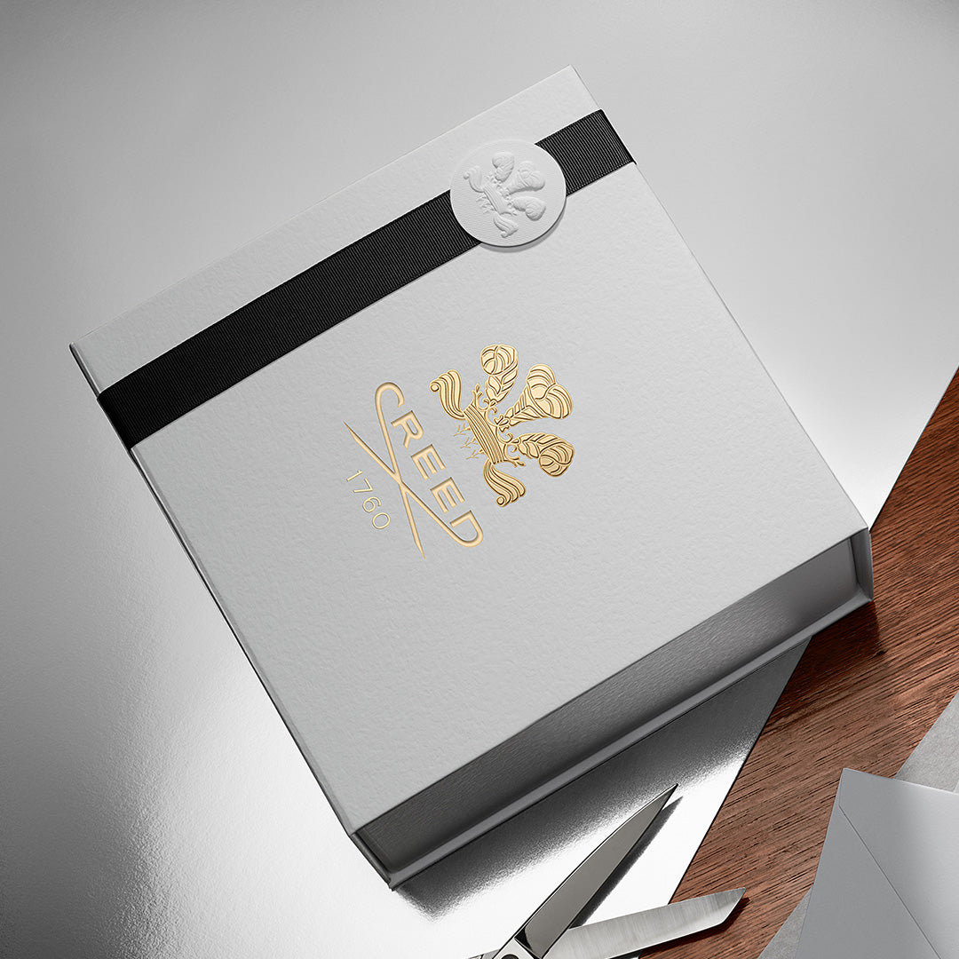 Silver box with gold logo and black ribbon on a gray surface with scissors.