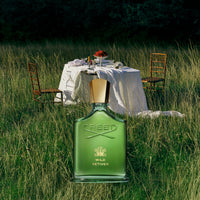 Green Creed Wild Vetiver perfume bottle in a grassy outdoor setting with a table and chairs.